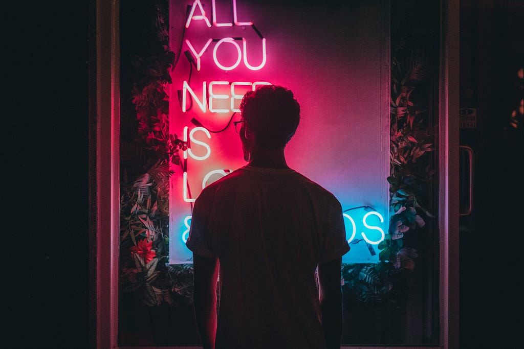 A man standing in front of neon lights ‘ALL YOU NEED IS LO…’