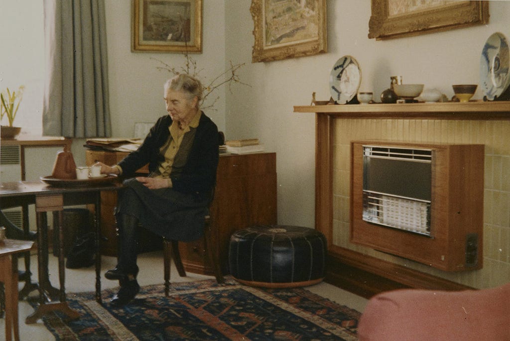 Colour photograph of Margaret Pilkington sat in her living room at home surrounded by paintings and pottery, 1969