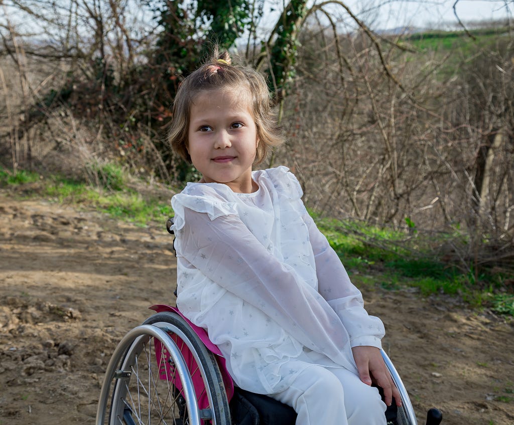 Picture of a young girl who is sitting on a wheel chair in park.