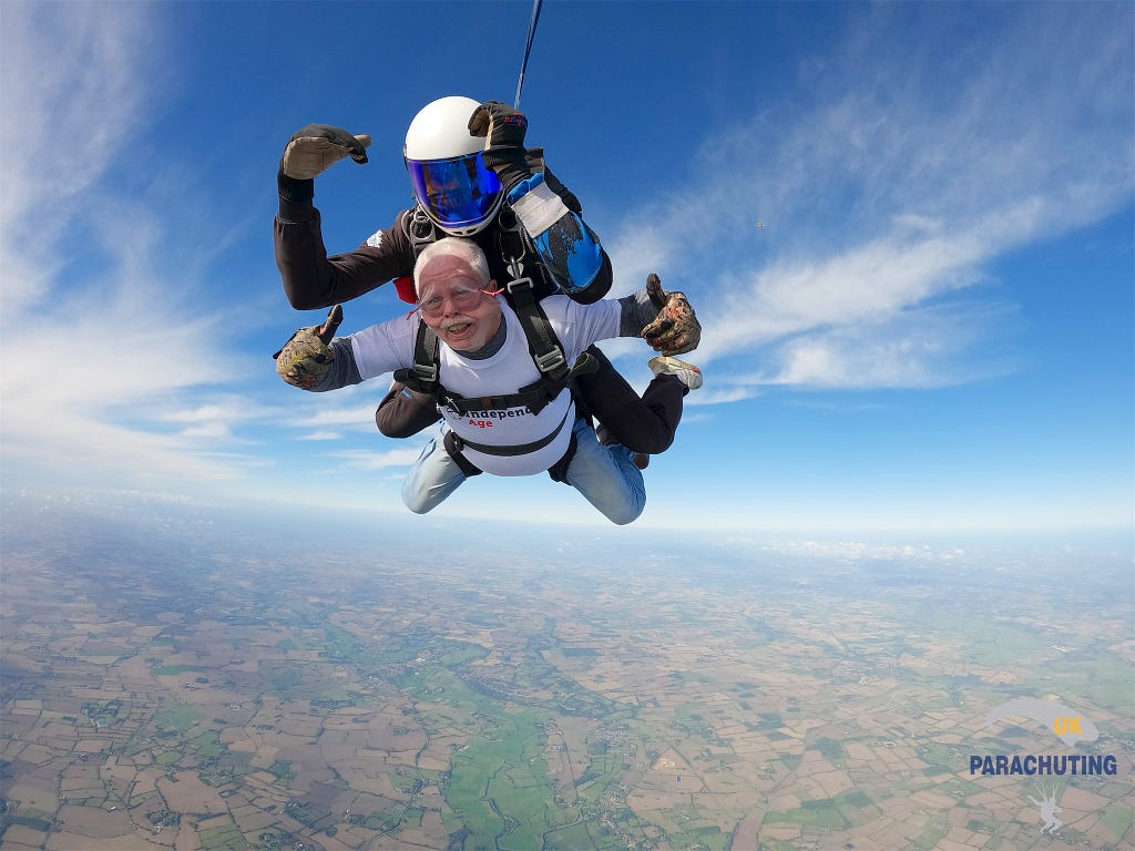 James, an older man, is attached to the front of another person as they skydive through the air. James holds his hands up, giving thumbs up to the camera.
