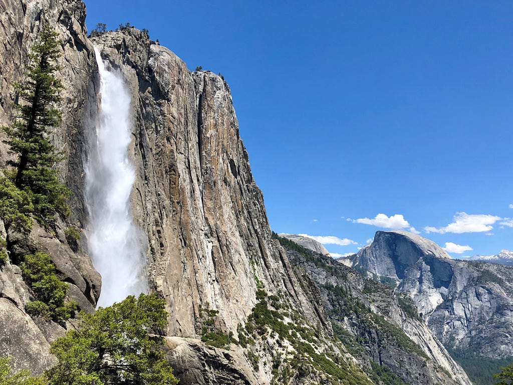 The raging water of Yosemite Falls tumbles over the sheer granite cliff. Half Dome towers in the background.