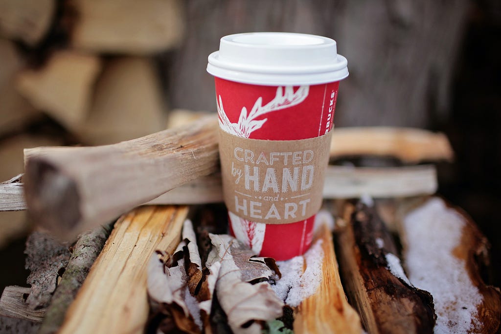 A red Starbucks cup sits on snowy branches. The coffee sleeve reads “Crafted by Hand and Heart.”