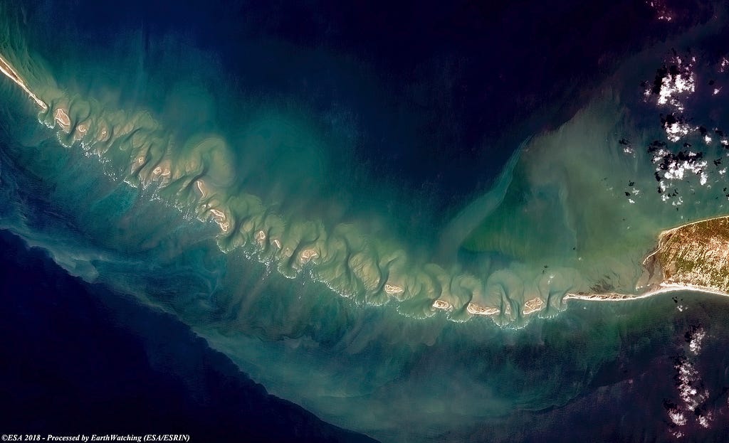 A high resolution satellite image shows water with a shallow area to the center and a rocky sandbar looking connection between the two landmasses.