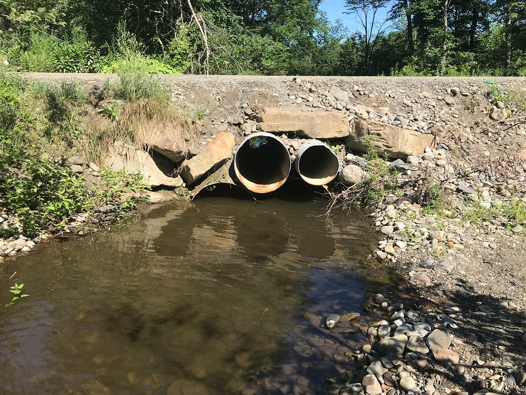 A roadway over a stream showing two large pipes just above the water