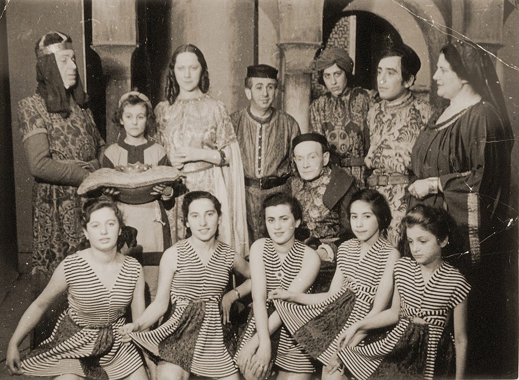 Thirteen people, a mix of male and female adults and children, in two rows standing indoors in front of a set used for a performance that has an arch and columns. The people in the back are dressed in elaborate royalty costumes. One actor holds a pillow with a crown prop on it. The five girls in front are wearing matching, horizontally striped dresses that they are holding out to their sides as if curtsying.