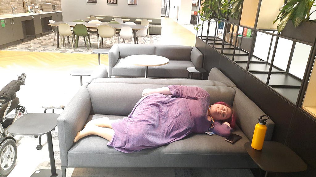 An office kitchen area with sofas. Claire is lying on a sofa, resting. Her wheelchair is to one side.