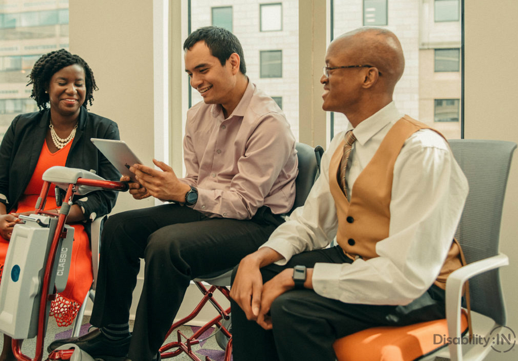 Stock photo of 3 people in a business setting, one woman, a man on a power wheelchair using an iPad, and another man on the r
