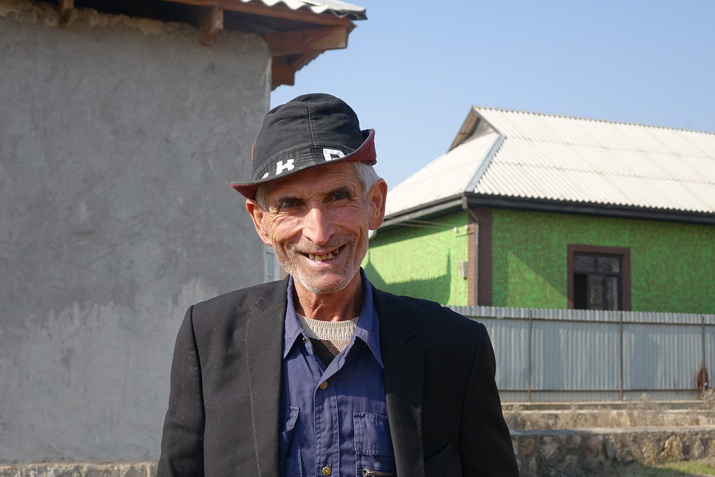 A older man with a wide smile and wearing a sharp black hat poses for a photo in front of his home.