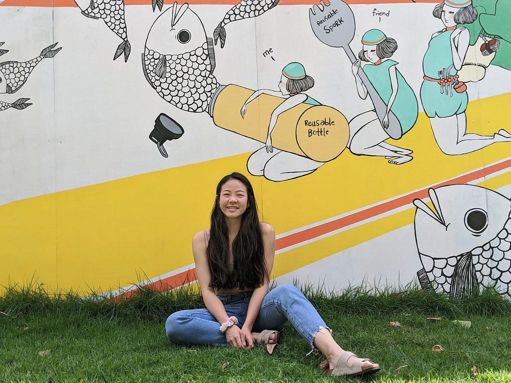 Western Biology graduate student LiAn Noonan sits on the grass in front of a wall with a brightly colored mural.