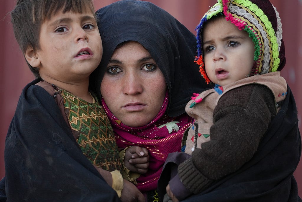 An Afghan woman displaced by war and drought near Herat, Afghanistan, holds her children as she waits for consultation outside a makeshift clinic at a sprawling settlement of mud-brick huts (Dec. 16, 2021). (AP Photo/Mstyslav Chernov)