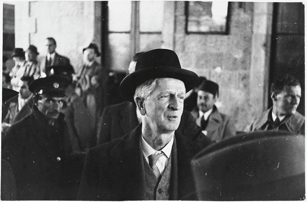 A man wearing an overcoat, vest, tie, and black hat looks off to the side of this black-and-white photograph. There are more men in business formal attire in the background. Over the man’s left shoulder, a man wearing a hat with a badge can be seen looking directly at the camera.
