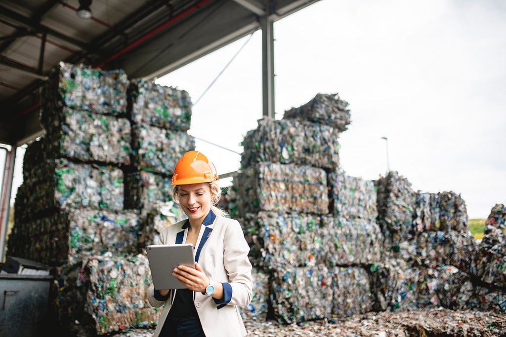 Woman working at a recycling facility wearing a white coat and hard hat writing on a clipboard