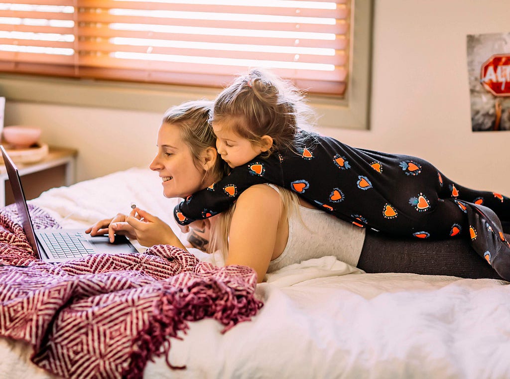 Inside a bedroom, a mother and child laying down on a bed using a laptop.