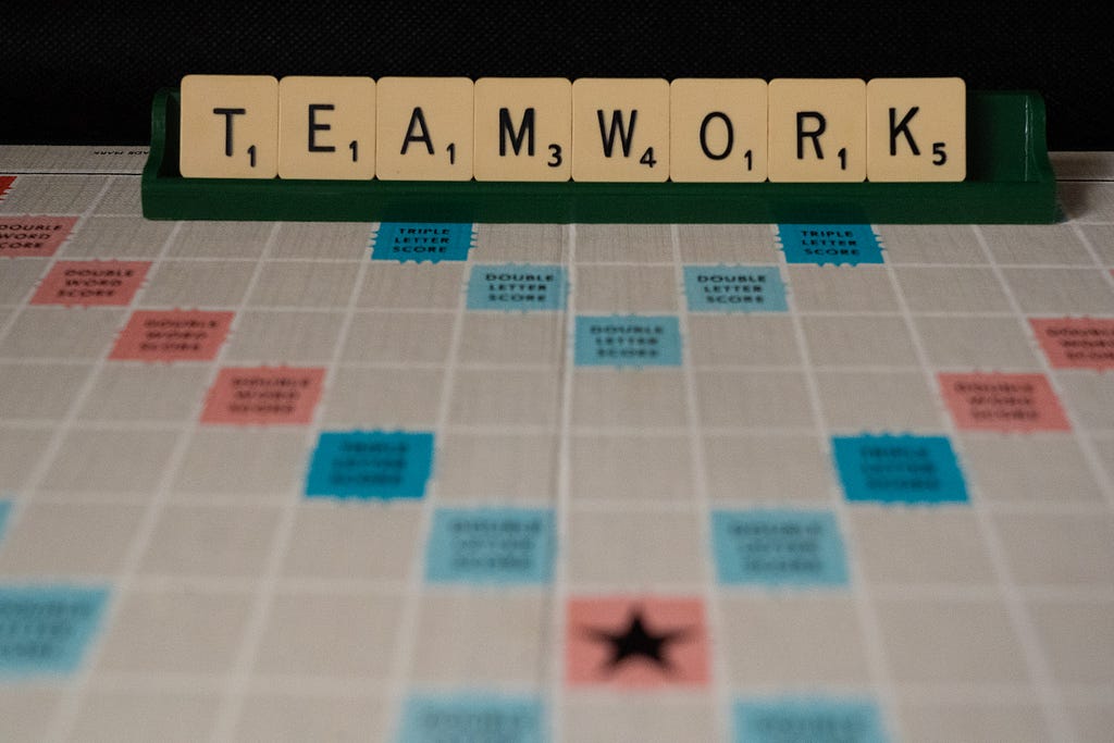 The word “TEAMWORK” is spelled out in Scrabble tiles on top of a game board.