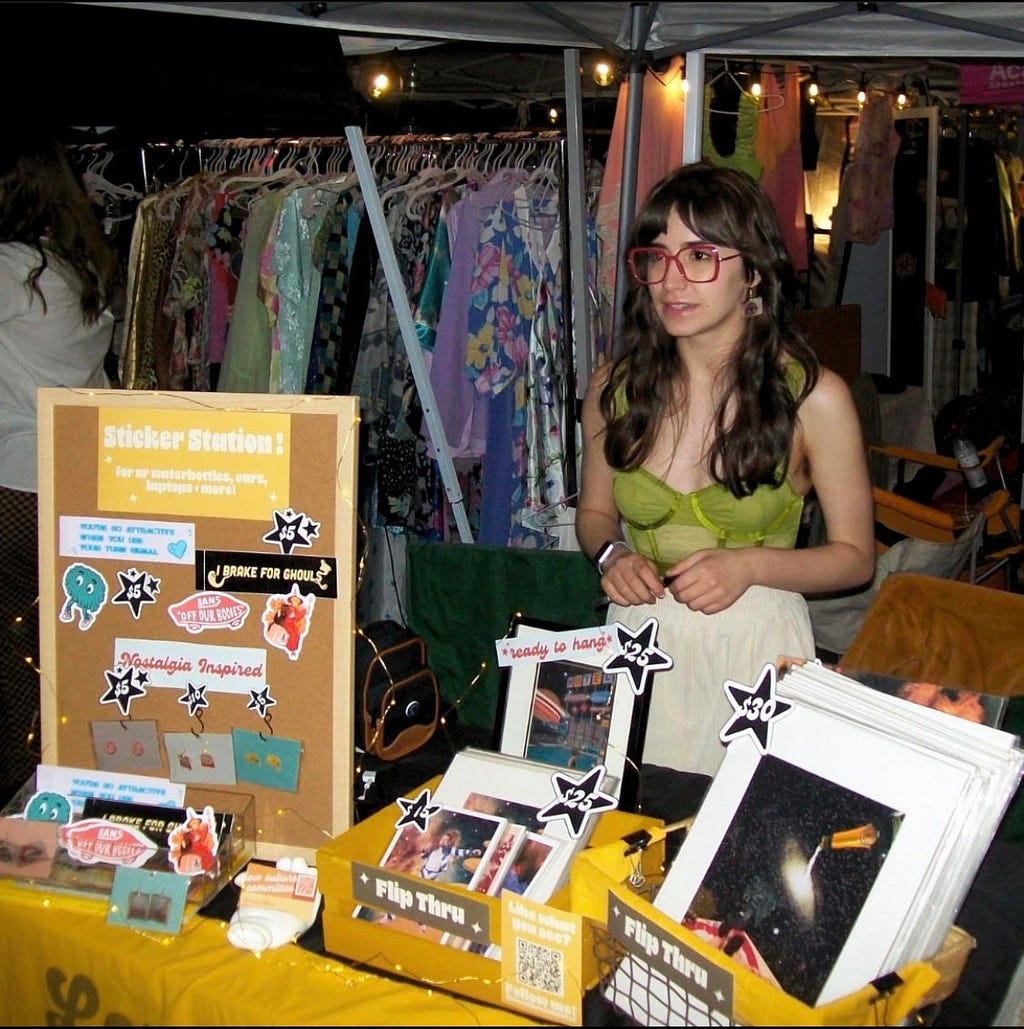 An image of Tori O’Campo at an art market standing behind a table full of stickers and art prints. She is wearing a green corset and white pants. Behind her is a rack full of color clothing.