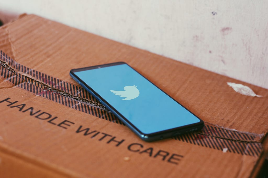 A phone with a blue screen and the twitter bird logo sits on a cardboard box that reads ‘handle with care’