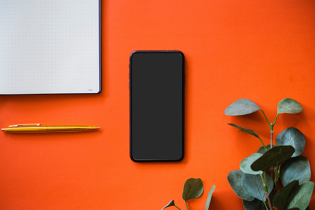 Orange background with black smartphone centered, surrounded by pen, plant leaves, notebook.