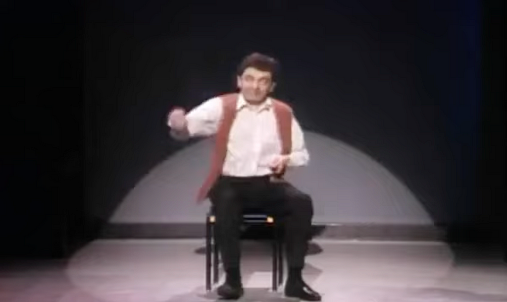 The image shows a man sitting on a chair on stage, performing in front of a dark background with a spotlight focused on him. He appears to be in motion, possibly acting or miming. The scene suggests a theatrical performance, with the man dressed casually in a vest and white shirt.