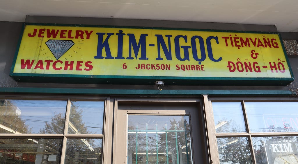 The Kim Ngoc storefront sign sits on top of the entrance, a yellow background with forest green trim. “Kim Ngoc” in all caps and navy blue texts sits at the center. To the left, the sign reads “jewelry” and “watches” in red, all caps text, the two words separated vertically by a diamond illustration. Underneath “Kim Ngoc” reads “6 Jackson Square” in the same red, all caps text. To the right are Vietnamese words in the same style.