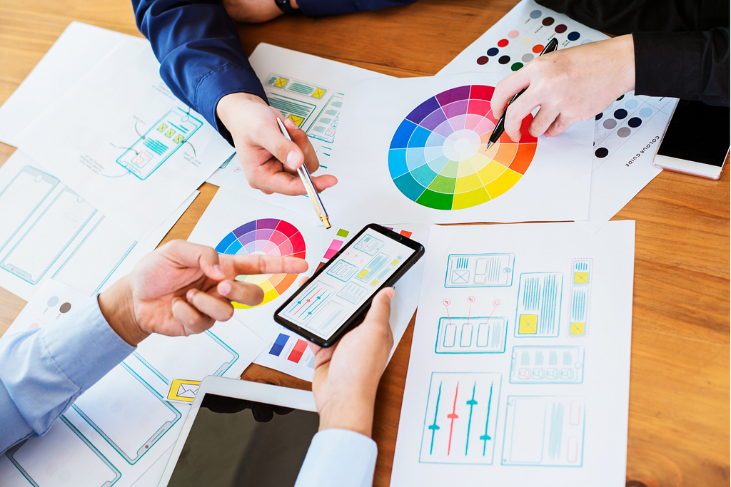 A group of people collaborating on a design project, discussing color schemes and interface layouts, with sketches and a color wheel spread across the table.