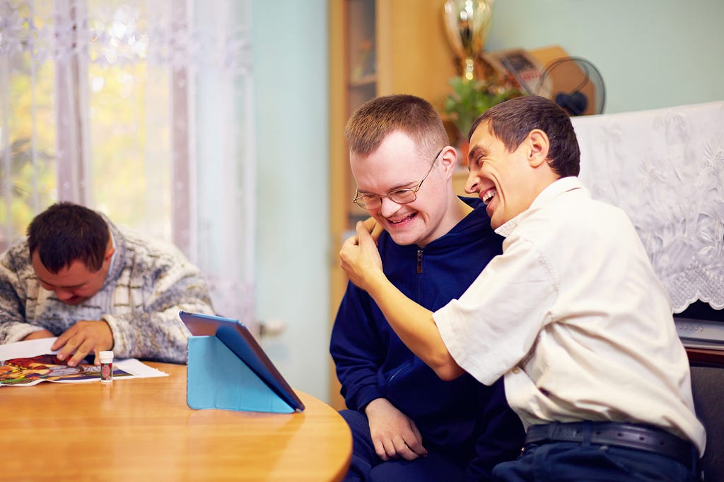 Two people sat at a table looking at a tablet. One of them is hugging the other and they are both laughing. In the background another person is sat at the table.