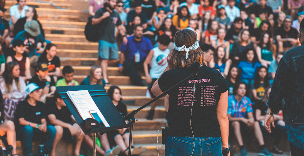 A woman standing in front of an auditorium and speaking, her back is turned to the camera, she’s wearing a t-shirt that has some dates on the back, potentially some tour