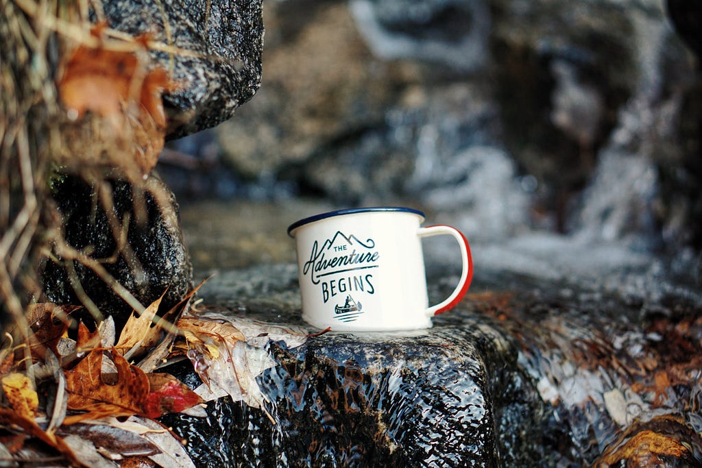A mug with The Adventure Begins sits outside on rocks