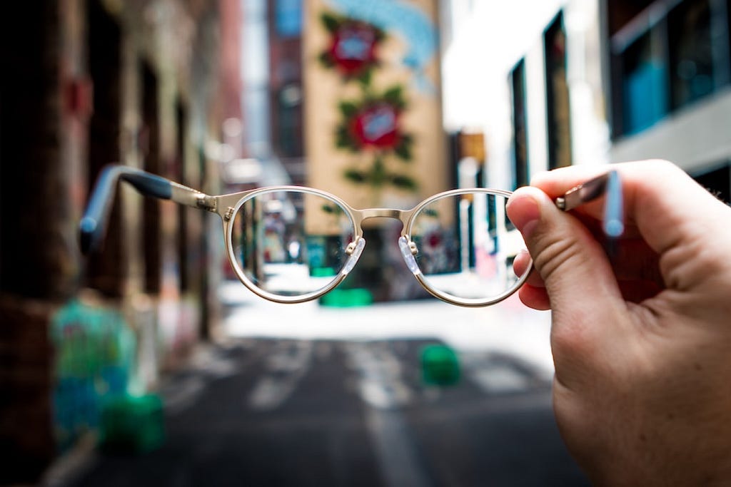 Image of hand holding glasses in front of a landscape of an alley. The landscape through the glasses is clear while the rest of the picture is blurry