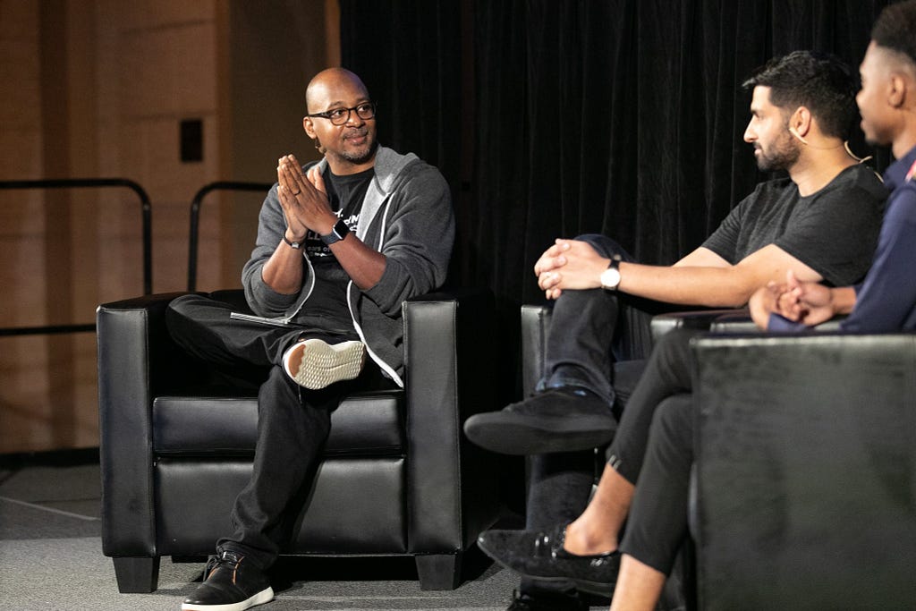 Wayne Sutton, a Black man wearing glasses and a hooded zip-up sweater, moderates a fireside chat on stage at Tech Inclusion.