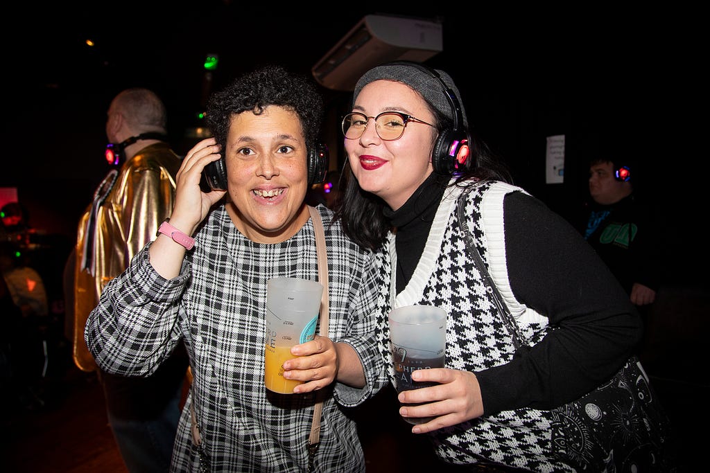 Two women smiling, holding drinks, dancing and wearing headphones in a dark night club