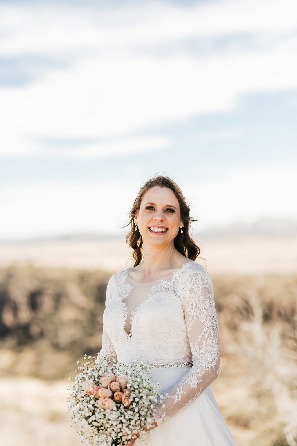 Picture of smiling bride (the author) in a white lace wedding dress with a faded scenery of a desert landscape behind her.