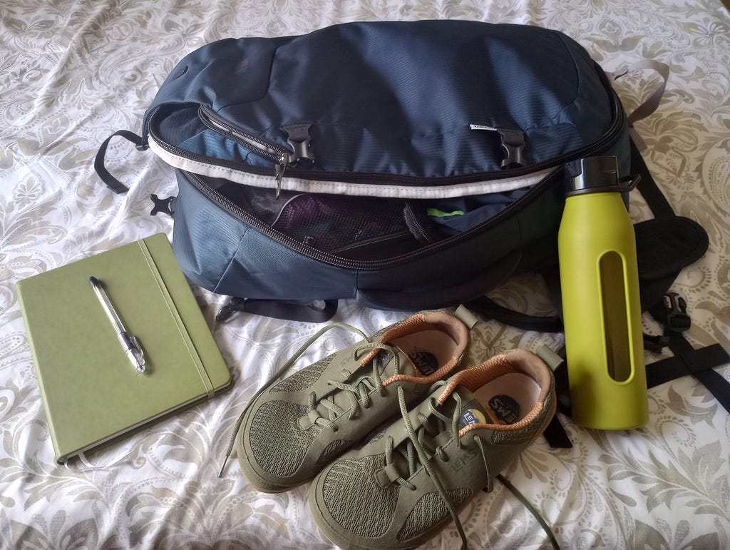 A dark blue backpack that opens like a clamshell is shown unzipped but with the lid still closed. A green journal with a pen, a pair of green shoes, and a green water bottle sit around it.