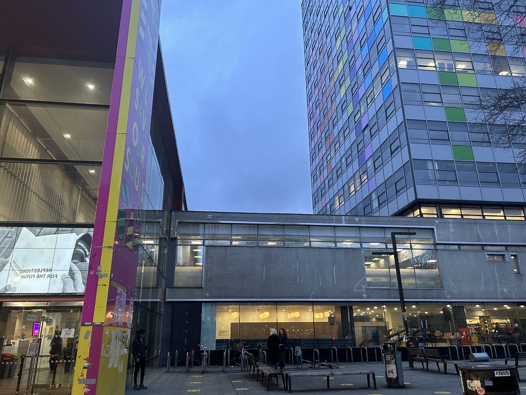 A picture of the front of the LCC UAL building, showing the entrance, the cafe from the outside and the tower block with students walking outside the bike racks.