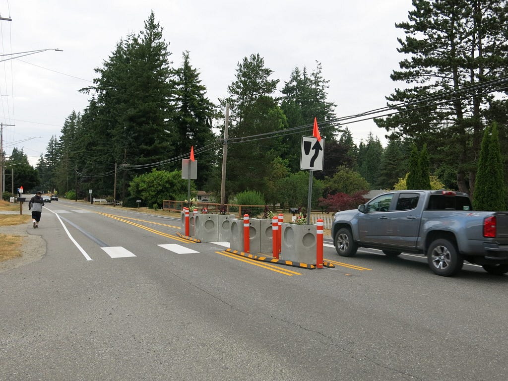 a pickup truck drives past the temporary crosswalk made of street signs and planters. A person can be seen on the left waiting to cross the street.