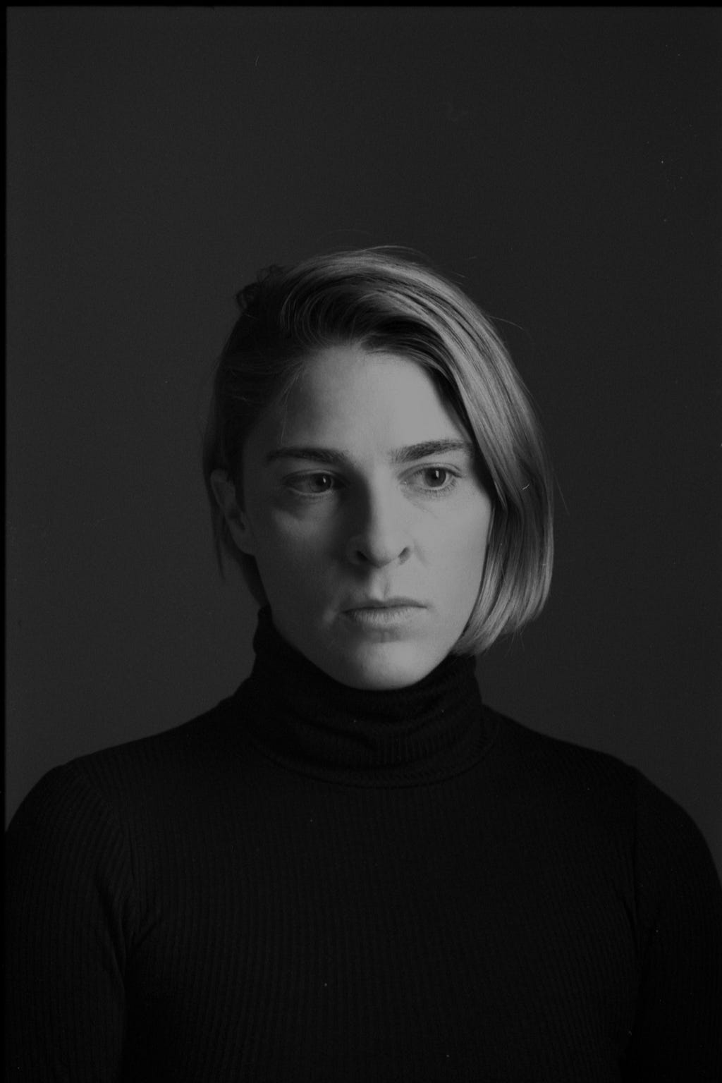 Black and white studio portrait of Alyssa she is looking off to her left. Her expression is serious. Her upper torso and head are centered in the frame. She wears a black turtleneck.