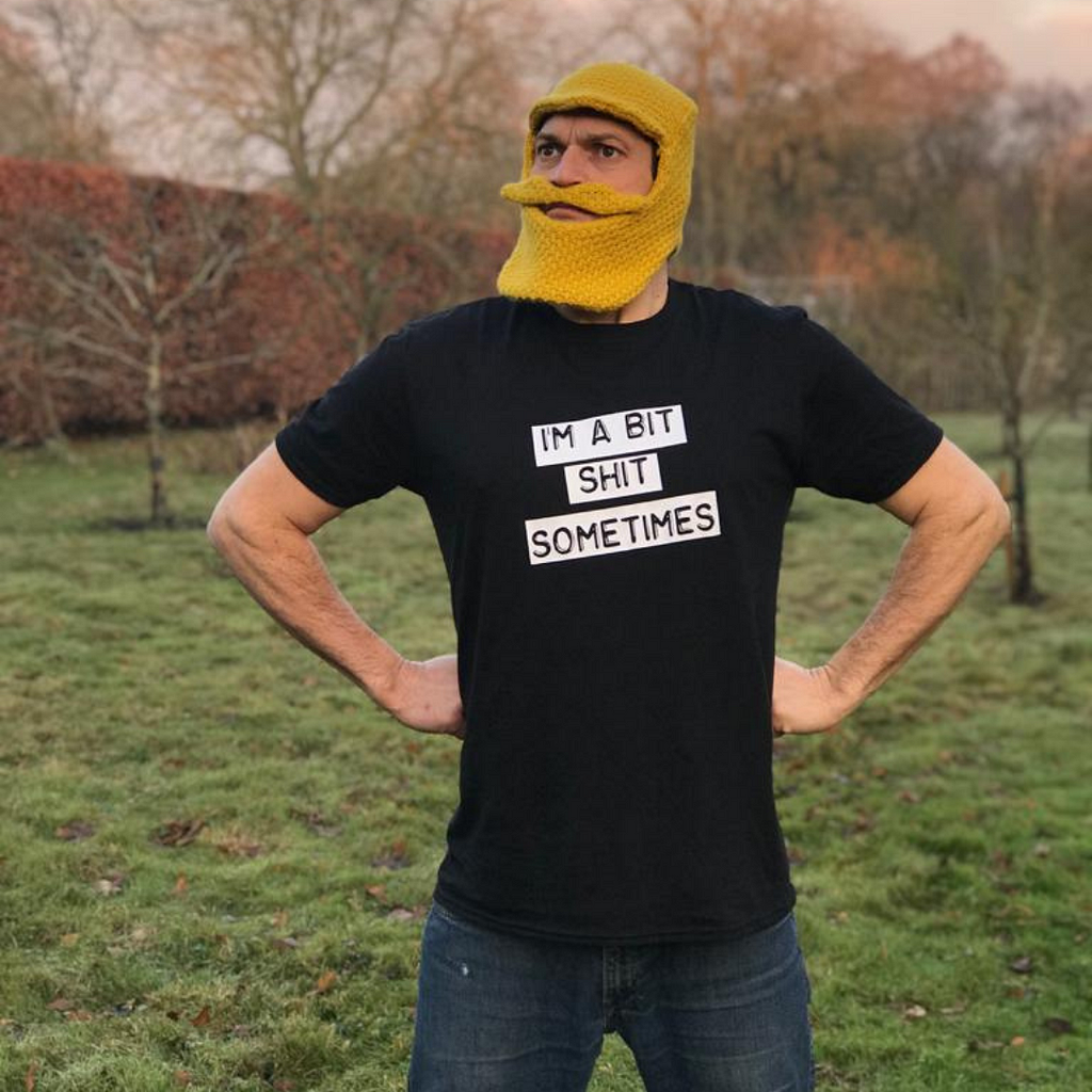 A man wearing a yellow knitted hat with a knitted beard poses with his arms on his hips. The t-shirt states “I’m a bit shit sometimes”.