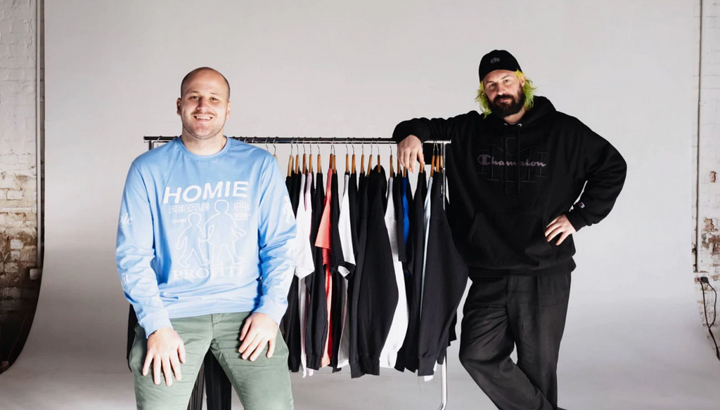 HoMie founders, Nick Pearce and Marcus Cook stand in front of a clothing rack full of t-shirt. Nick is wearing a blue shirt with ‘Homie’ on the front. Marcus is wearing a black jumper and cap.