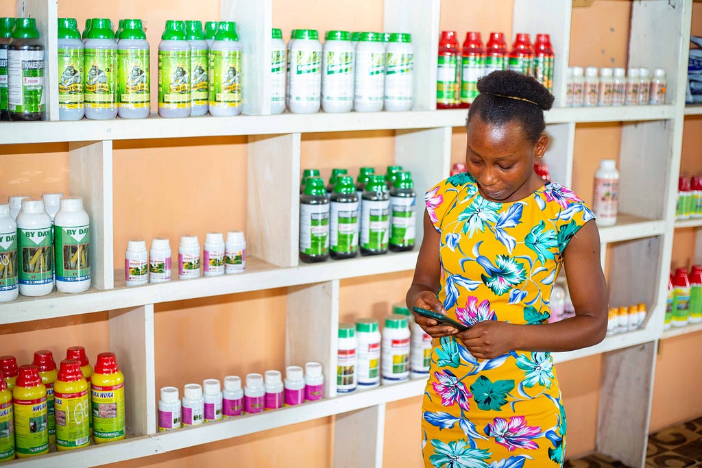 A woman looks down at her mobile phone while standing next to shelves lined with bottles.