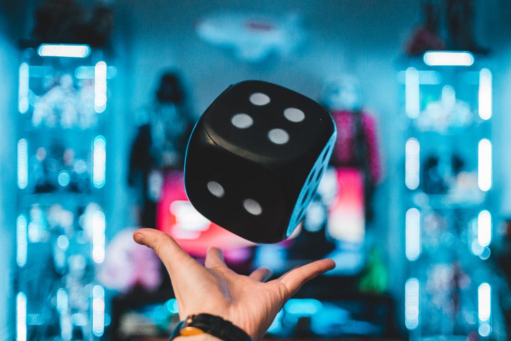 A hand beneath an oversized die. The die is black with white spots.
