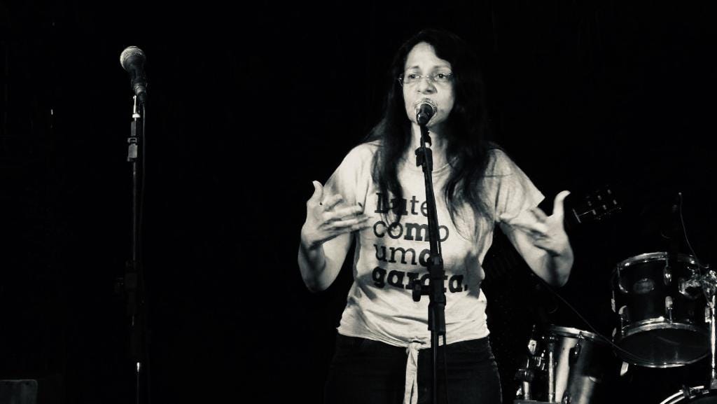 A imagem mostra Dora Gadelha, uma mulher branca, de óculos e cabelos pretos. Ela usa uma camisa branca com a frase “Lute como uma garota” e gesticula com os braços. Está falando em um microfone, com uma bateria musical à sua direita e somente um fundo preto a suas costas. A foto está em preto e branco.