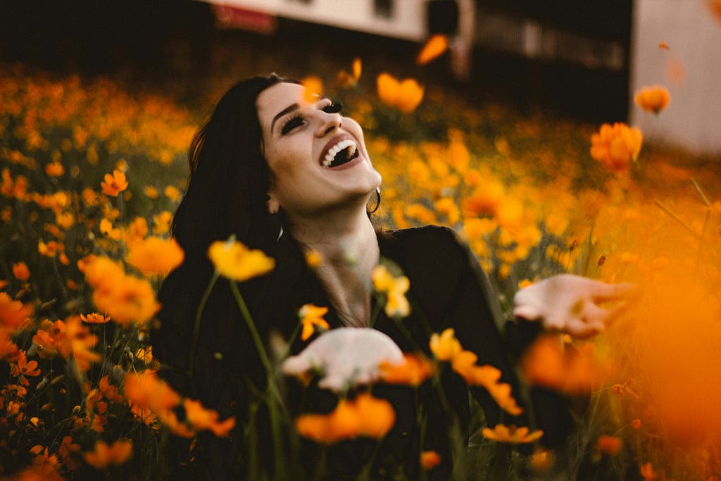 A woman with an overjoyed expression sits in a field of marigolds, some of which appear to be drifting towards sky