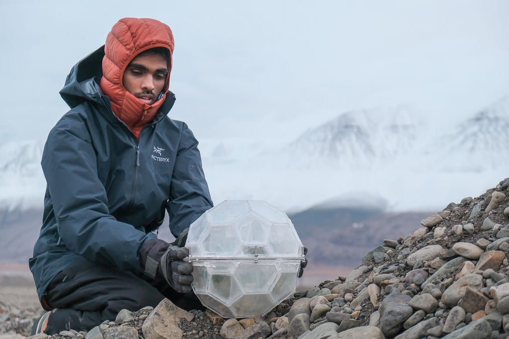 Somayajulu Dhulipala wearing a red hooded jacket and black wind breaker, holding the AgriThrive plant habitat—a clear white orb made of hexagonal and pentagonal prisms—on a bed of rocks. White mountains misty in the background.