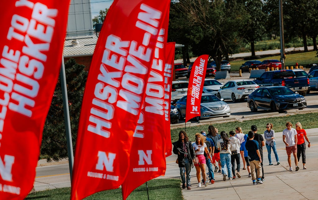 People walk along a sidewalk lined with flags that read “Husker Move-In”