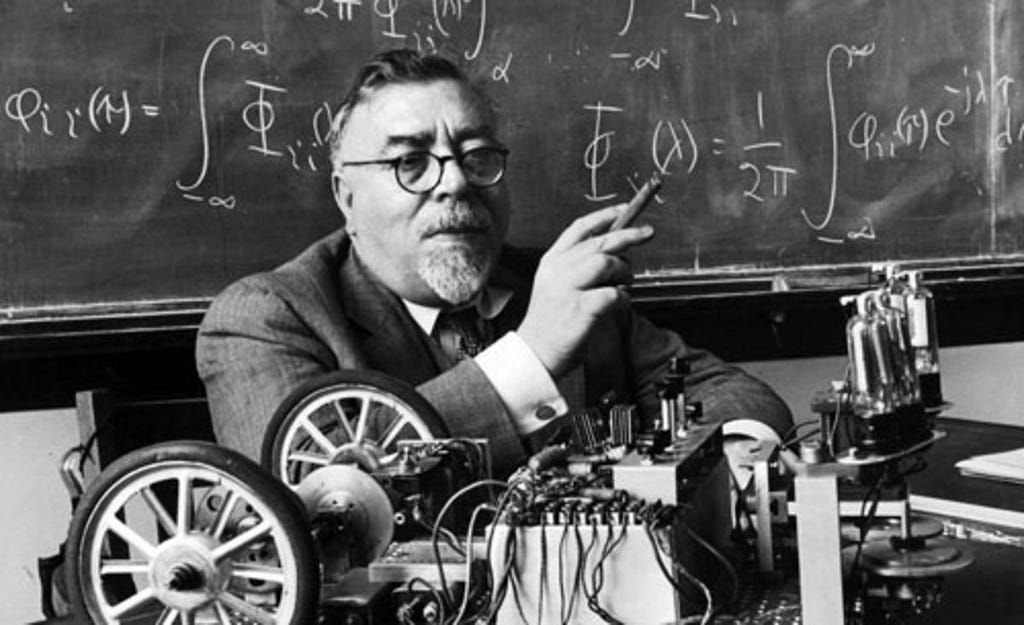 Cyberneticist Norbert Wiener sitting in front of a blackboard with the wheeled robot, Palomilla