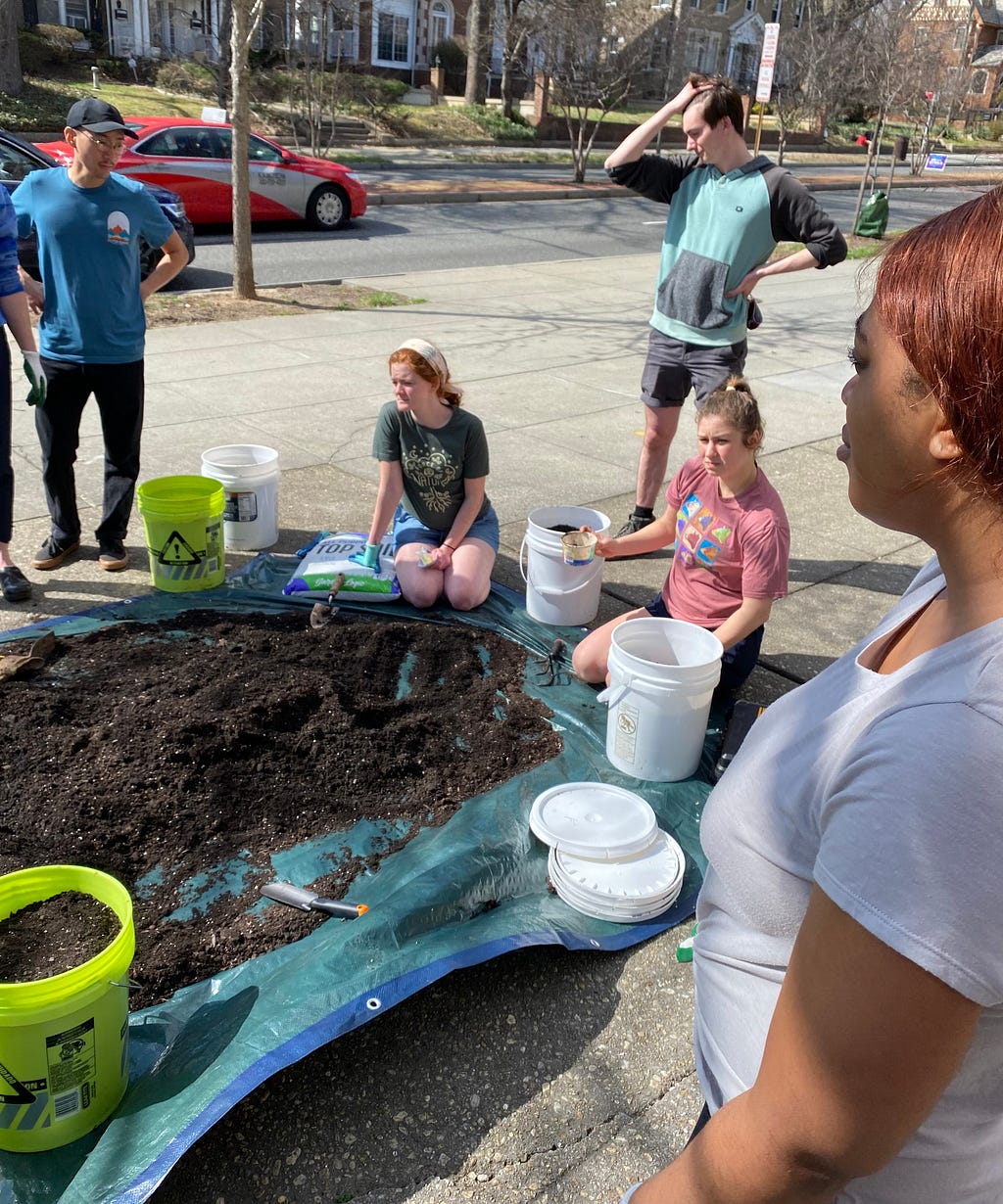 Five people standing around a tarp with soil and buckets in order to plant vegetables for community garden.