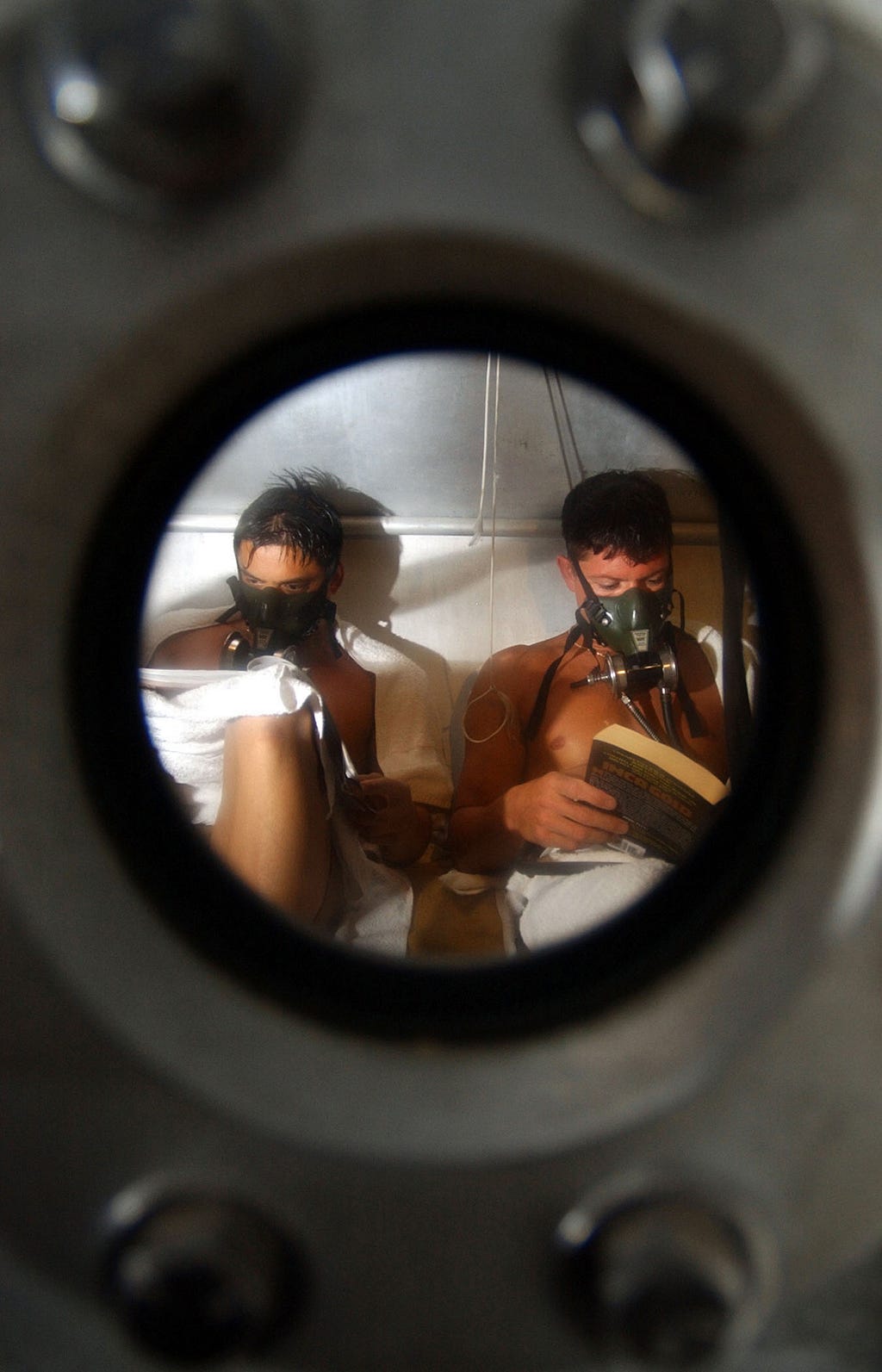 Through the view of a porthole, two young men are seen reading while wearing ocygen masks.