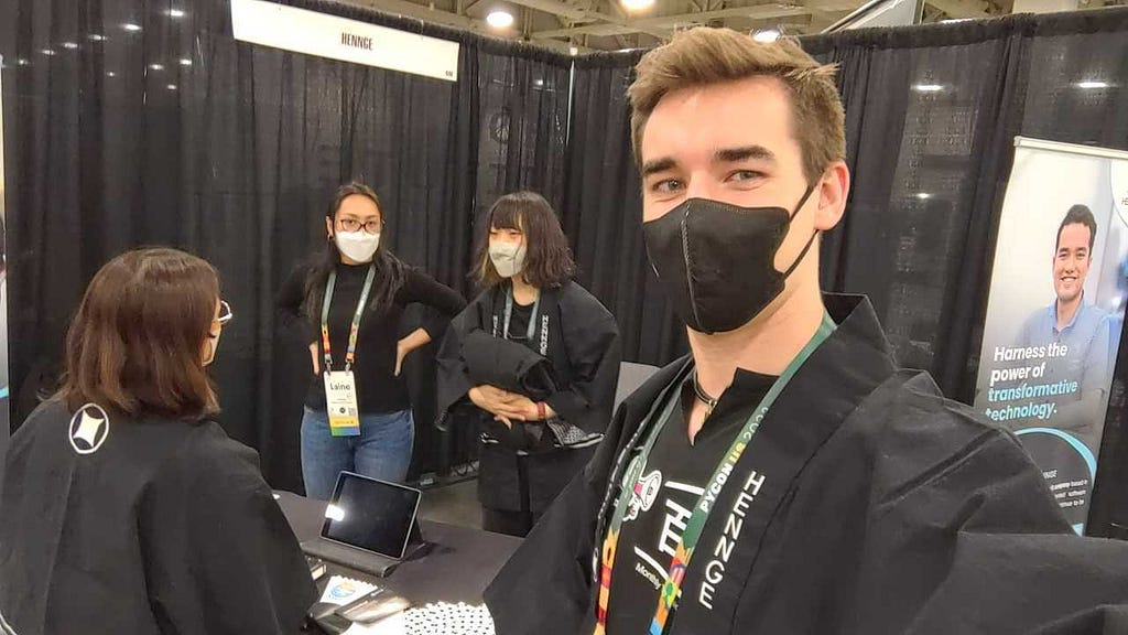 A selfie from the author of himself at HENNGE’s booth at PyCon 2023. He’s looking into the camera and his coworkers are in a small group to the side.