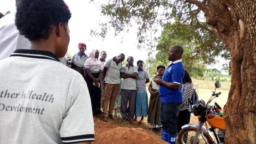 A man stands under a tree and next to a motorcycle, with a group of people circled up around him.