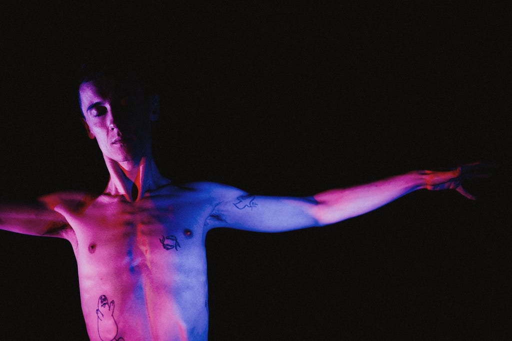 A young man is shirtless with arms out-stretched. He is against a black background and is lit by purple and blue light