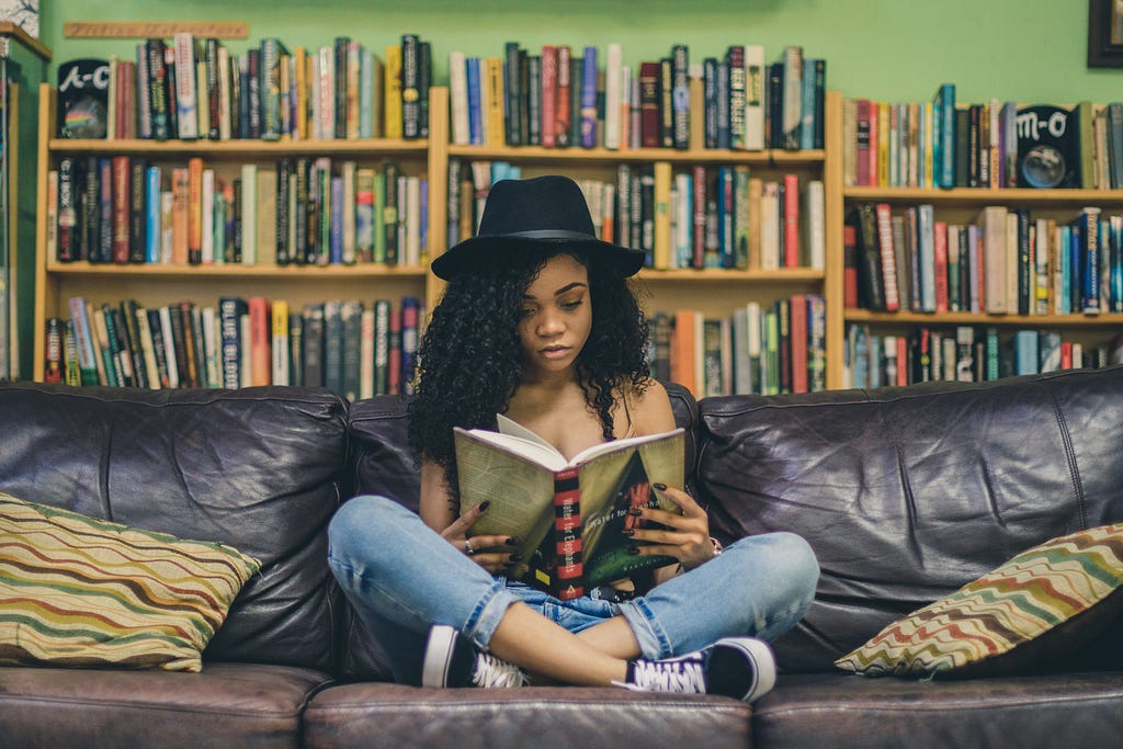 A woman, on a couch, reading a book with a bookshelf behind her